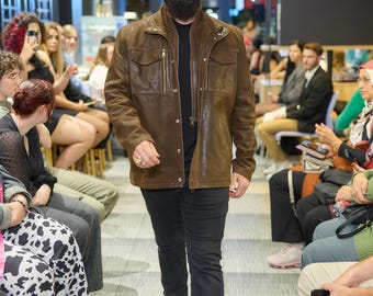 May include: A man wearing a brown leather jacket, black jeans, and yellow boots walks down a runway. He is also wearing a black cap and sunglasses. The background shows an audience seated on both sides.