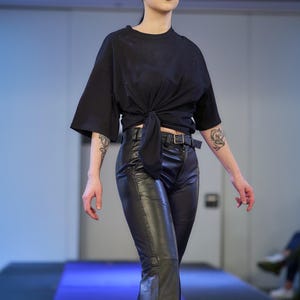 May include: A black, short-sleeved top tied at the waist, paired with black leather pants. The pants have a flared leg design and a belt. The model is on a blue runway.