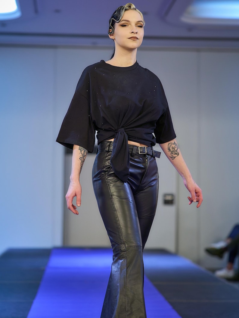 May include: A black, short-sleeved top tied at the waist, paired with black leather pants. The pants have a flared leg design and a belt. The model is on a blue runway.