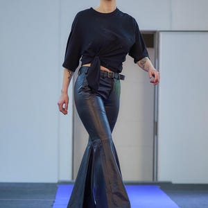 May include: A person wearing a black t-shirt tied at the waist, paired with black flared leather pants and a black belt. The person is wearing white shoes and walking on a blue carpet.