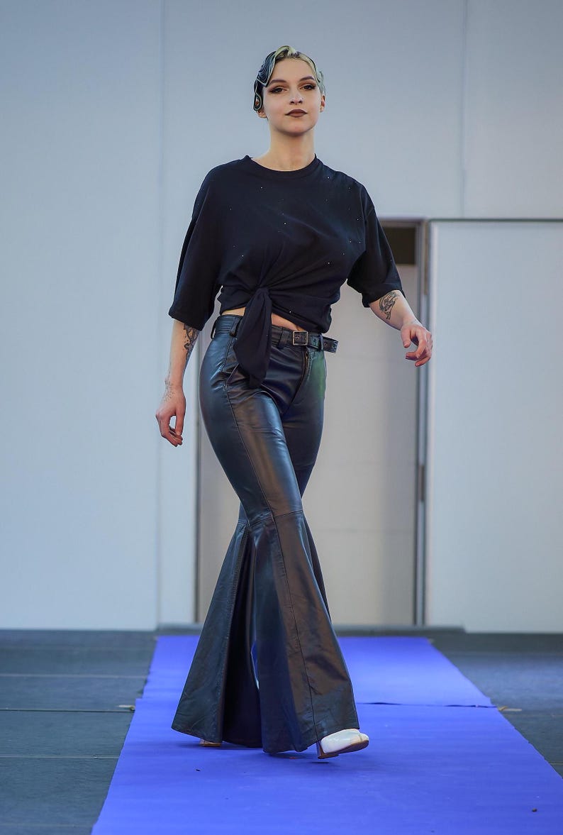 May include: A person wearing a black t-shirt tied at the waist, paired with black flared leather pants and a black belt. The person is wearing white shoes and walking on a blue carpet.