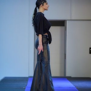 May include: A black, tied-front top and flared leather pants. The outfit is worn with white shoes. The model is standing on a blue runway.