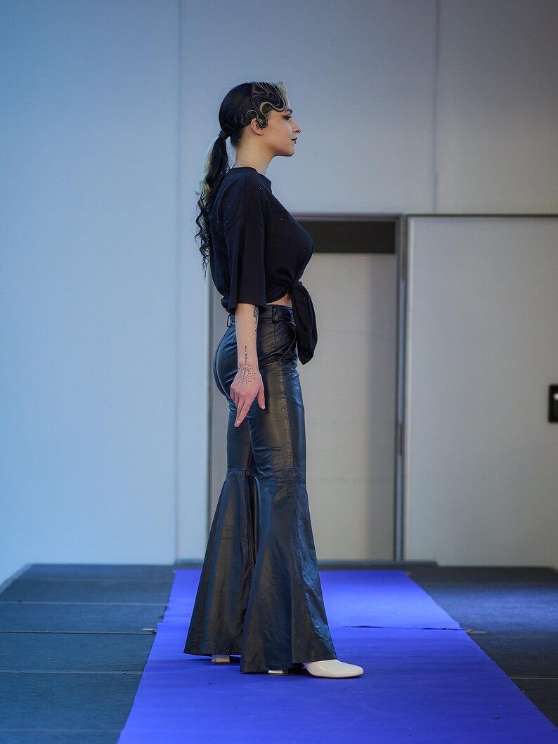 May include: A black, tied-front top and flared leather pants. The outfit is worn with white shoes. The model is standing on a blue runway.