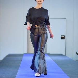 May include: A model wearing a black knotted top, black leather pants with a flared leg, and white shoes walks on a blue runway. The pants have a belt with a silver buckle.