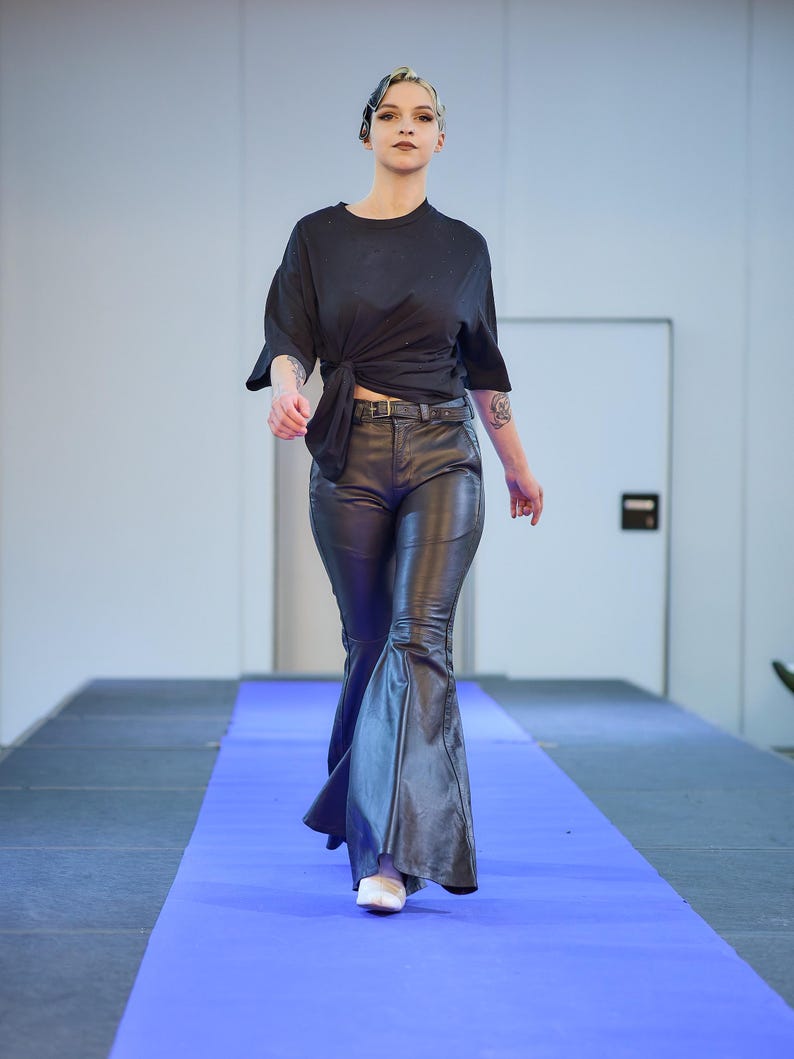 May include: A model wearing a black knotted top, black leather pants with a flared leg, and white shoes walks on a blue runway. The pants have a belt with a silver buckle.