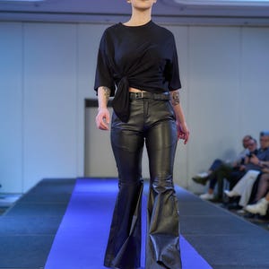 May include: A model walks a runway wearing a black top tied at the waist, black leather pants, and white shoes. The pants have a flared leg design. The model is on a blue runway.