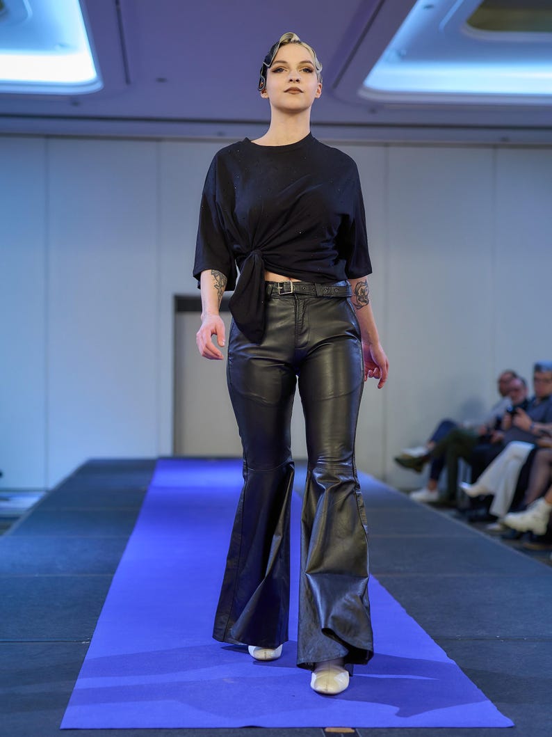 May include: A model walks a runway wearing a black top tied at the waist, black leather pants, and white shoes. The pants have a flared leg design. The model is on a blue runway.
