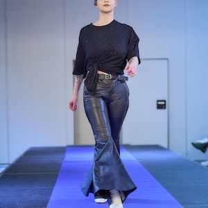 May include: A model walks a runway wearing a black knotted t-shirt, black leather pants with a flared leg, and white boots. The runway is covered in a blue carpet.