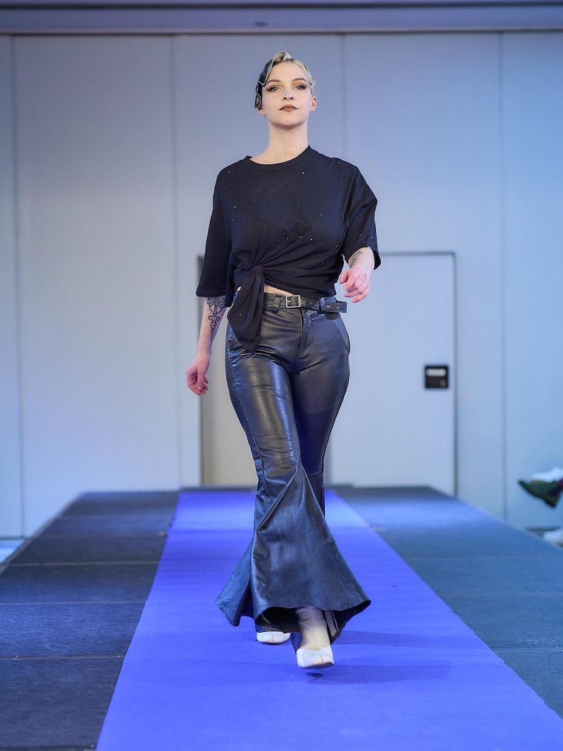 May include: A model walks a runway wearing a black knotted t-shirt, black leather pants with a flared leg, and white boots. The runway is covered in a blue carpet.