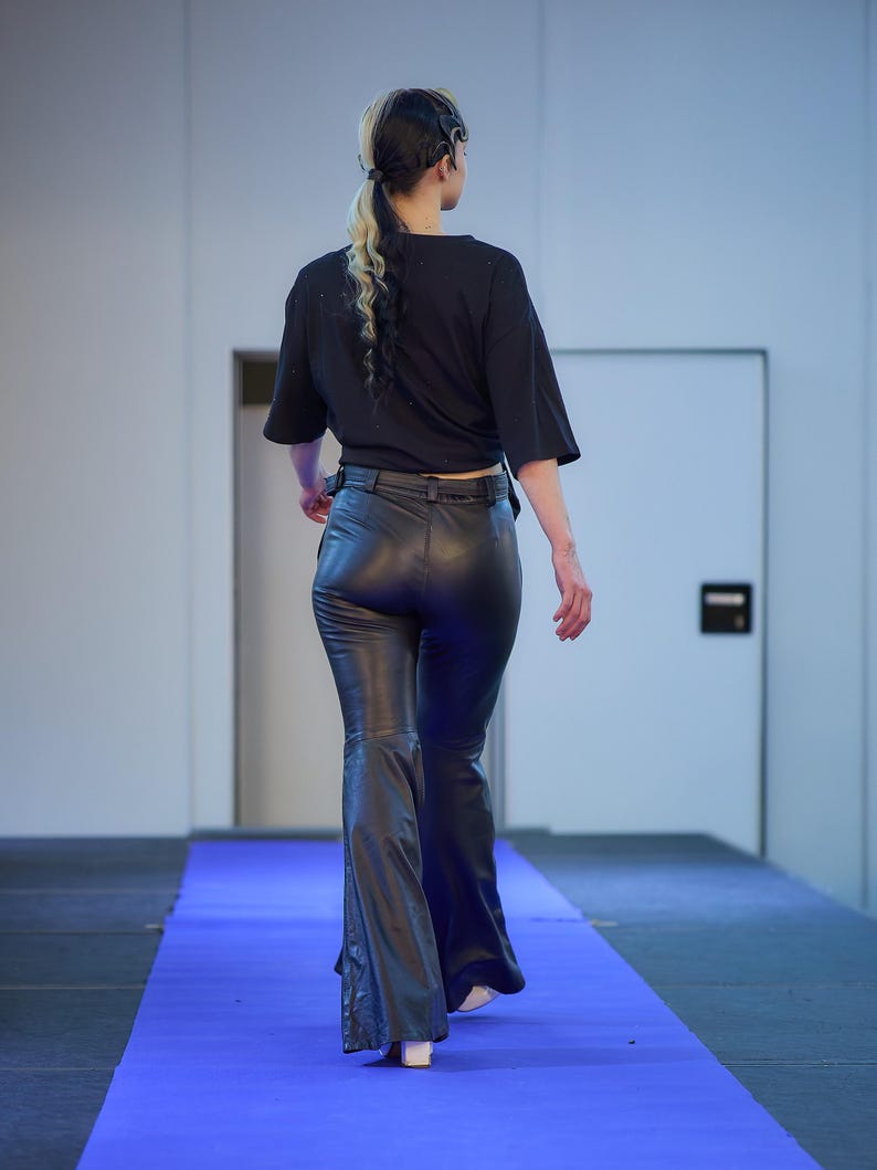 May include: A person wearing a black short-sleeved shirt and black leather flared pants walks on a blue carpet. The outfit is paired with white shoes. The person has blonde and black hair.
