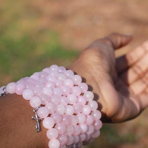 May include: A stack of light pink beaded bracelets with a silver cross charm. The bracelets are made of round, polished beads. The image shows the bracelets on a person's arm, with the hand open.
