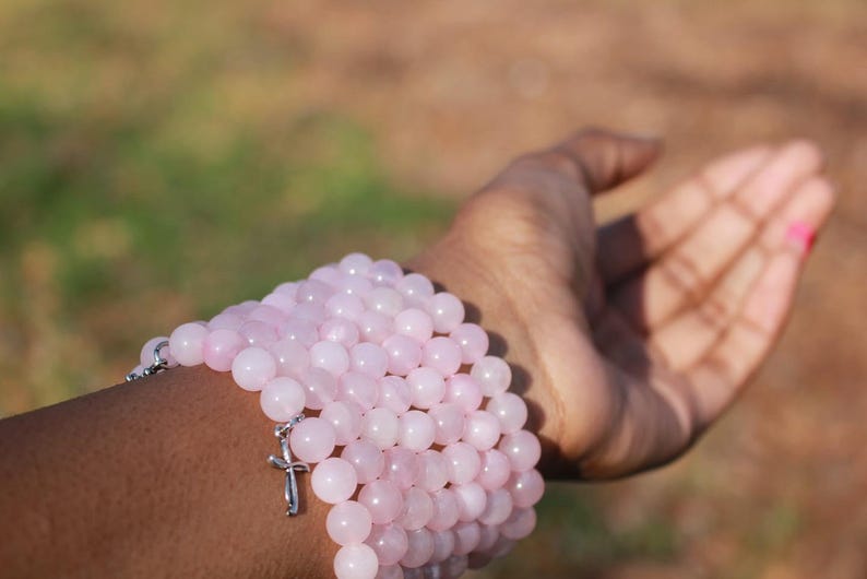 May include: A stack of light pink beaded bracelets with a silver cross charm. The bracelets are made of round, polished beads. The image shows the bracelets on a person's arm, with the hand open.