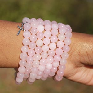 May include: A stack of light pink beaded bracelets with a silver cross charm. The bracelets are made of round, polished beads. The bracelets are worn on a person's wrist.