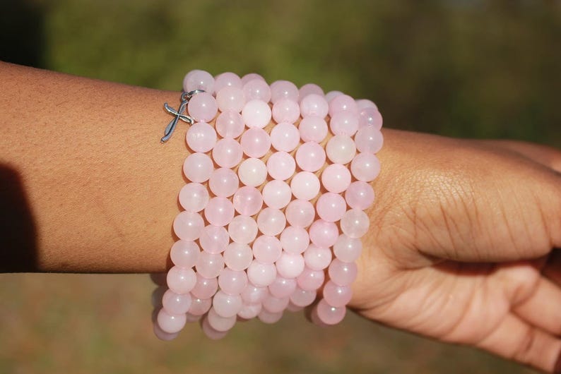May include: A stack of light pink beaded bracelets with a silver cross charm. The bracelets are made of round, polished beads. The bracelets are worn on a person's wrist.
