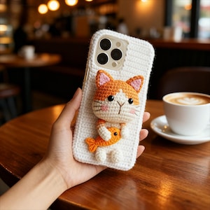 Może przedstawiać: Białe, szydełkowe etui na telefon z 3D pomarańczowo-białym kotem trzymającym pomarańczową rybę. Kot ma różowe akcenty na uszach i policzkach. Etui jest przeznaczone do smartfona.