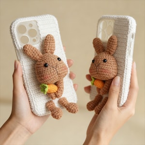 Może przedstawiać: Dwa białe, szydełkowe etui na telefon, każde ozdobione 3D szydełkowym brązowym królikiem trzymającym marchewkę. Królik ma czarne oczy i różowe policzki. Etui są trzymane w dłoniach.