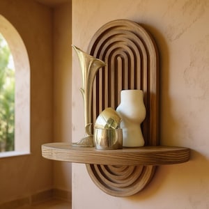 May include: A wooden shelf with a carved arch design, holding decorative items. A gold-colored trumpet-shaped vase, two gold-colored bowls, and a white, curvy vase are displayed. The shelf is mounted on a peach-colored wall, creating a warm, inviting aesthetic.