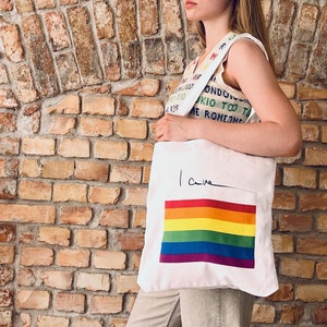 Op de afbeelding: Een witte tote bag met een regenboogvlag en het woord "love" in het zwart. De tas heeft een regenboogband.