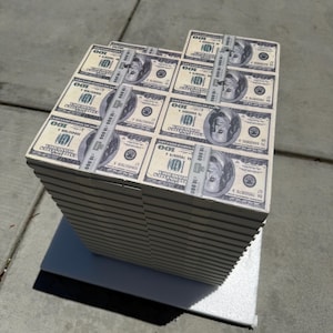 May include: A large stack of United States hundred-dollar bills, secured with paper bands. Each bill features Benjamin Franklin and the number 100. The stack is placed on a white surface.