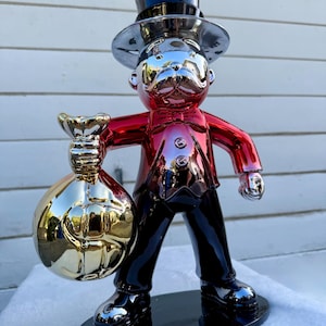 May include: A reflective, metallic figurine of a character wearing a black top hat, a red jacket, and black trousers. The figure holds a large, shiny gold-toned money bag in one hand and stands on a black oval base with a polished finish.