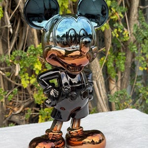 Puede incluir: Una figura de Mickey Mouse de pie con un acabado metálico reflectante en negro y bronce. El personaje aparece con las manos en la cintura, vistiendo pantalones cortos con dos botones y zapatos grandes y redondeados. La superficie es brillante.