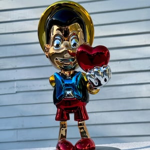 May include: A reflective, metallic-finish figurine of a character wearing a yellow hat, a blue bowtie, and red shorts. The figure has a glossy, mirror-like surface and is holding a large, shiny red heart in one hand, standing on a black base.