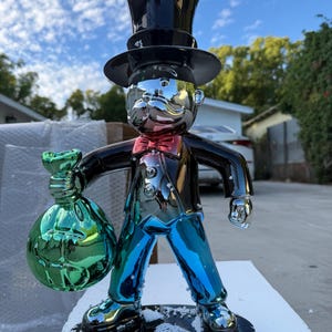 Puede incluir: Una figura reflectante y multicolor de un personaje con sombrero de copa, pajarita roja y traje, que sostiene una gran bolsa de dinero verde. La figura tiene un acabado metálico tipo espejo con tonos azules, negros y plateados sobre una base.