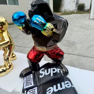 Puede incluir: Una escultura de gorila negro en posición de boxeo, con guantes de boxeo azules y dorados y pantalones cortos rojos. La escultura se encuentra sobre un par de guantes de boxeo negros con la palabra "Supreme" en blanco. Los guantes tienen el logo "Everlast".