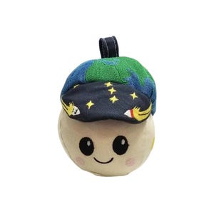 Puede incluir: Un peluche con forma de hongo sonriente con una gorra azul y verde. La gorra está decorada con un mapa del mundo y estrellas. El hongo tiene una cara feliz con mejillas rosadas y un lazo negro para colgar.