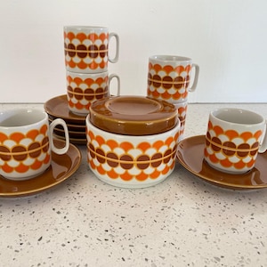 May include: A vintage ceramic coffee set featuring mugs, saucers, and a lidded sugar bowl. The set has a white base with a repeating pattern of orange and brown semi-circles. The saucers and sugar bowl lid are brown.