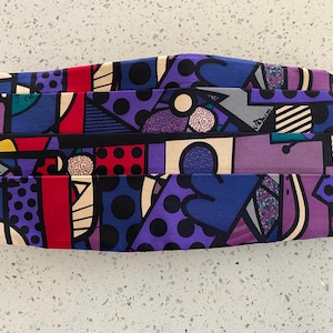 May include: A pleated cummerbund featuring a vibrant, multi-colored abstract pattern with geometric shapes, circles, and dots in shades of purple, blue, red, black, and cream. The fabric displays a repeating graphic design with distinct color blocks.