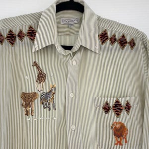 May include: A button-down shirt with vertical stripes in beige and brown. The shirt features embroidered safari animals, including a giraffe, zebra, elephant, and lion. Decorative diamond shapes with a zebra print pattern are also embroidered on the shirt.