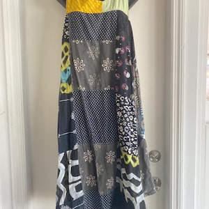 May include: A patchwork dress with a variety of patterns and colors, including yellow, pink, green, black, and white. The dress is sleeveless and hangs on a black mannequin. The dress features polka dots, floral designs, and abstract shapes.