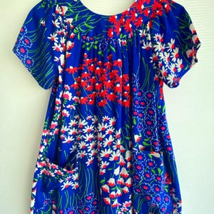 May include: A vibrant blue dress featuring a repeating floral pattern in shades of red, white, and green. The garment has short sleeves, a rounded neckline, and two front patch pockets integrated into the floral design, hanging on a black hanger.