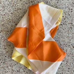 May include: Folded square silk scarf with a bold geometric pattern. The scarf features alternating stripes of orange, white, and yellow. The fabric has a slight sheen, suggesting a luxurious material. The scarf is folded, showcasing the color block design.