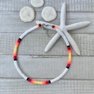 May include: A white beaded necklace with a gradient of red, orange, and yellow beads. The necklace is on a wooden surface with a starfish and seashells.