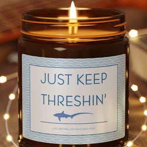 May include: A brown glass candle with a white label that reads "JUST KEEP THRESHIN'" and a small blue shark graphic. The candle is lit and sitting on a wooden coaster.
