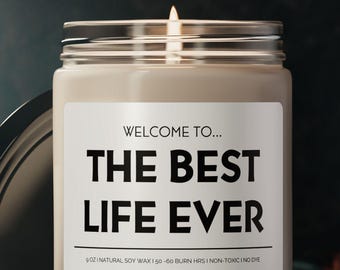 May include: A lit candle in a clear glass jar with a silver lid. The label reads "WELCOME TO... THE BEST LIFE EVER" in bold black text. The candle is on a dark surface with the lid slightly ajar.