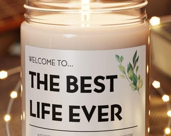 May include: A lit candle in a glass jar with the text "WELCOME TO... THE BEST LIFE EVER" on a white label with green leaf accents. The candle sits on a round wooden coaster, surrounded by string lights.