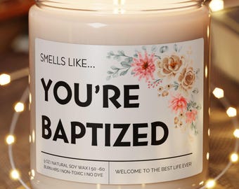 May include: A lit candle in a clear glass jar with the text "YOU'RE BAPTIZED" on a label with floral accents. The candle sits on a round wooden coaster, surrounded by string lights. A guitar and book are in the background.