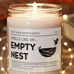 May include: A lit candle in a glass jar with a label that reads "EMPTY NEST." The candle sits on a wooden coaster, surrounded by string lights. The label also says "Quiet Home Aromatherapy" and "Smells like an..."