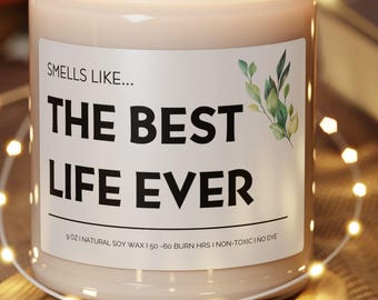May include: A lit candle in a glass jar with the text "THE BEST LIFE EVER" on a white label. The candle sits on a round wooden coaster, surrounded by string lights. A guitar and a book are in the background.