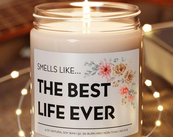 May include: A lit candle in a clear glass jar with a label that reads "Smells like... The Best Life Ever." The candle sits on a round wooden coaster. A string of fairy lights and a guitar are in the background.