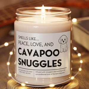 Puede incluir: Una vela encendida en un tarro de cristal con una etiqueta que dice "Smells Like... Peace, Love, and Cavapoo Snuggles". La vela está sobre un posavasos redondo de madera, rodeada de luces de cadena. Una guitarra y un libro están en el fondo.