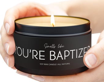 May include: A lit black candle in a round tin. The candle has a gold rim and the text "Smells like YOU'RE BAPTIZED" in white. The bottom of the tin reads "SOY WAX CANDLE | ALL NATURAL".