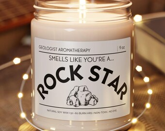 May include: A lit, scented candle in a clear glass jar with a label that reads "Smells Like You're a Rock Star." The candle sits on a small wooden coaster, with string lights and a guitar in the background. The candle is 9 oz.