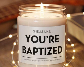 May include: A lit candle in a clear glass jar with a white label that reads "SMELLS LIKE... YOU'RE BAPTIZED." The candle sits on a small, round wooden coaster. A string of fairy lights and a guitar are in the background.