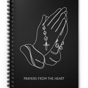 May include: Black spiral-bound notebook with a white line drawing of praying hands and a rosary. The words "Prayers From The Heart" are printed in white at the bottom of the cover.