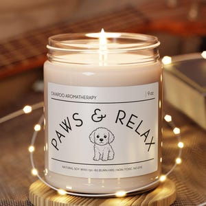 Puede incluir: Una vela perfumada encendida en un tarro de cristal transparente, con la etiqueta "PAWS & RELAX" con una ilustración de perro. La vela está sobre un pequeño posavasos de madera. La vela es de 255 gramos.