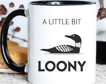 May include: White ceramic mug with a black handle and rim. The mug features the text "A LITTLE BIT LOONY" with a black illustration of a loon bird. The mug is on a light-coloured surface.